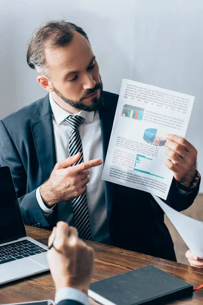 Geschäftsmann zeigt auf Papier mit Diagrammen in der Nähe von Laptop, Investor und Kollege im verschwommenen Vordergrund — Stockfoto