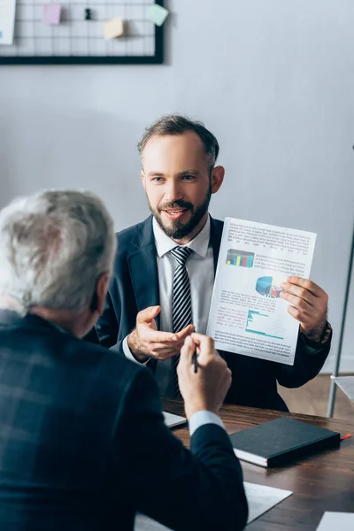 Geschäftsmann zeigt Papier mit Grafiken in der Nähe eines reifen Investors auf verschwommenem Vordergrund — Stockfoto