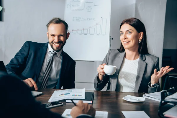 Cheerful businesspeople with coffee talking with advisor on blurred foreground near papers on table — Stock Photo