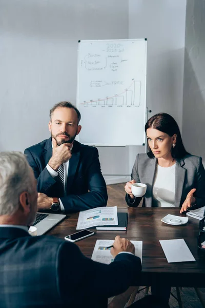 Femme d'affaires avec tasse de café parler près de collègue et conseiller sur l'avant-plan floue au bureau — Photo de stock