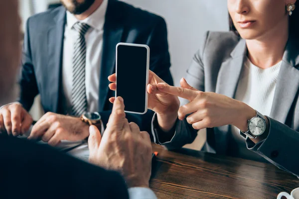 Vista recortada de la mujer de negocios apuntando al teléfono inteligente con pantalla en blanco cerca de colega e inversor en primer plano borroso - foto de stock