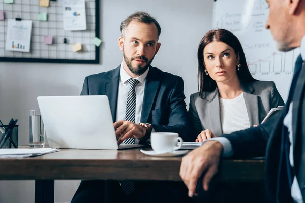 Businesspeople with laptop looking at mature investor on blurred foreground in office — Stock Photo