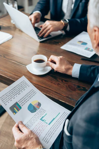 Paper with charts and coffee in hands of businessman near partner with laptop on blurred background — Stock Photo