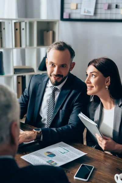 Smiling businesspeople with documents looking at investor on blurred foreground in office — Stock Photo