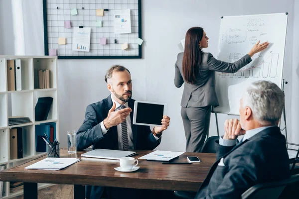 Geschäftsmann zeigt auf digitales Tablet neben Kollege, Flipchart und Investor im verschwommenen Vordergrund im Büro — Stockfoto