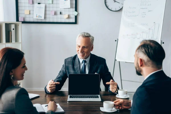 Smiling investor pointing at laptop with blank screen near business people with coffee on blurred foreground — Stock Photo