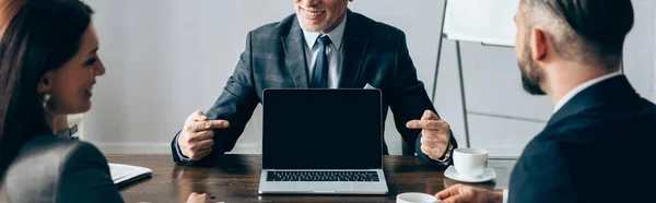 Lächelnder Berater zeigt auf Laptop in der Nähe von Geschäftspartnern mit Kaffee im verschwommenen Vordergrund, Banner — Stockfoto