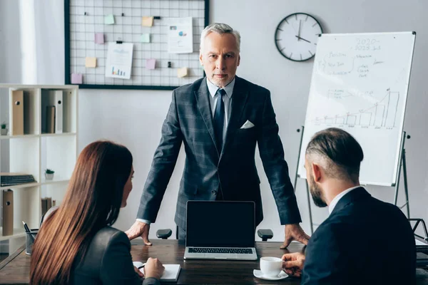 Reifer Investor blickt auf Kamera in Laptopnähe mit leerem Bildschirm und Geschäftspartnern im verschwommenen Vordergrund — Stockfoto