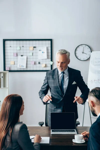 Lächelnder Berater zeigt auf Laptop neben Geschäftsfrau mit Notizbuch und Kollegin mit Tasse Kaffee im verschwommenen Vordergrund — Stockfoto