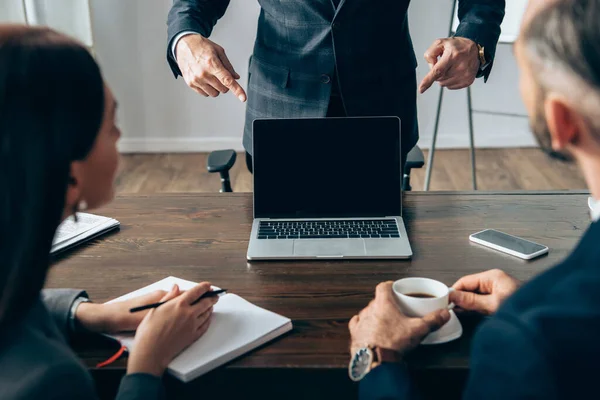 Investor pointing with fingers at laptop with blank screen near colleagues with coffee and notebook on blurred foreground — Stock Photo