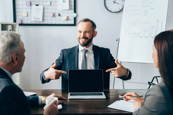 Investidor sorrindo apontando para laptop perto de empresários com café e notebook em primeiro plano turvo durante a consulta — Fotografia de Stock