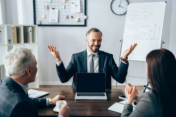 Cheerful businessman looking at partners near laptop with blank screen and papers in office — Stock Photo