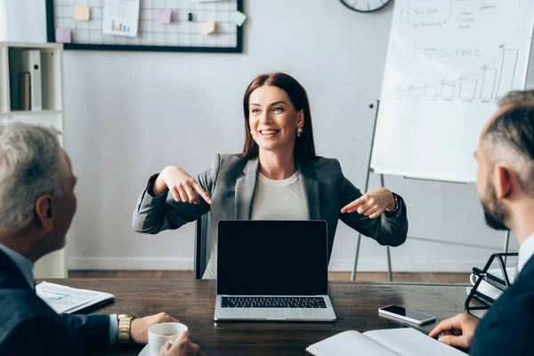 Lächelnde Geschäftsfrau zeigt auf Laptop mit leerem Bildschirm in der Nähe von Partnern mit Notebook und Kaffee im verschwommenen Vordergrund — Stockfoto