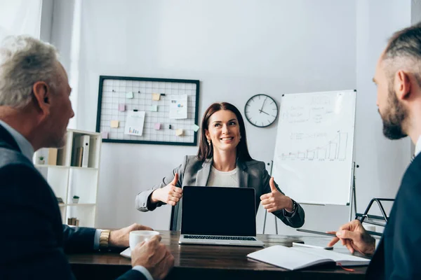 Smiling businesswoman showing thumbs up near partners on blurred foreground and laptop with blank screen — Stock Photo