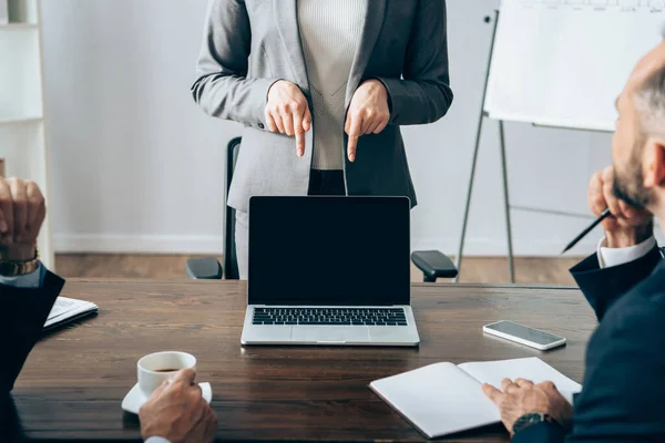 Empresaria señalando con los dedos a la computadora portátil con pantalla en blanco cerca de socios con cuaderno y café en primer plano borroso en la oficina - foto de stock