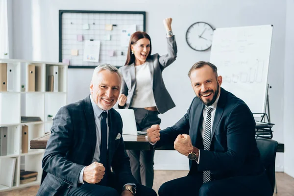 Cheerful businessmen showing yeah gesture near colleague on blurred background in office — Stock Photo