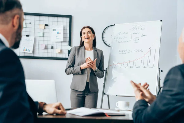 Donna d'affari sorridente in piedi vicino flipchart vicino a partner commerciali in primo piano sfocato — Foto stock