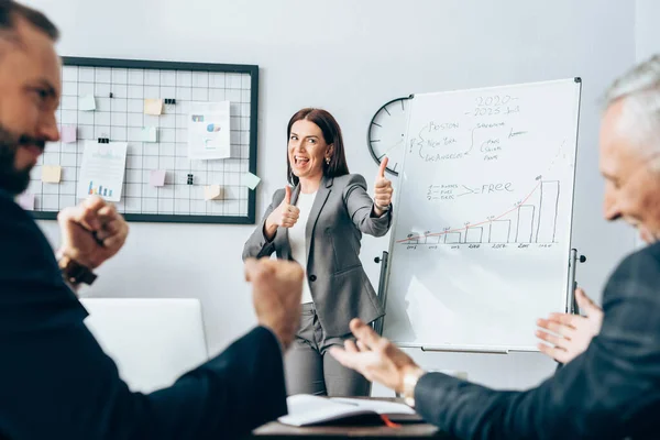 Cheerful businesswoman showing like gesture near flipchart and colleagues on blurred foreground — Stock Photo