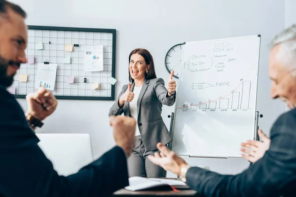 Mulher de negócios alegre mostrando como gesto perto flipchart e colegas em primeiro plano desfocado — Fotografia de Stock