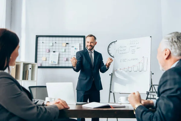 Lächelnder Geschäftsmann zeigt Ja-Geste in der Nähe von Flipchart mit Grafiken und Kollegen im verschwommenen Vordergrund — Stockfoto