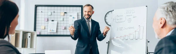 Fröhlicher Geschäftsmann zeigt Yah-Geste in der Nähe von Flipchart und Partnern auf verschwommenem Vordergrund im Büro, Banner — Stockfoto