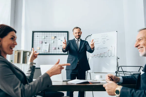 Smiling businessman pointing with fingers at colleagues on blurred foreground near flipchart in office — Stock Photo