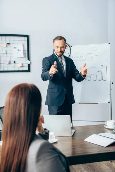 Uomo d'affari sorridente che punta con le dita vicino flipchart e collega in primo piano sfocato in ufficio — Foto stock