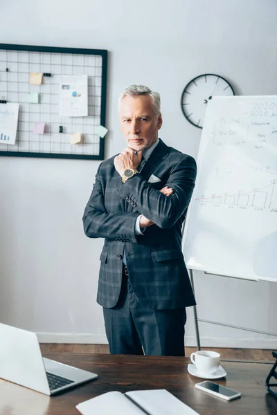 Selbstbewusster Geschäftsmann blickt auf Kamera in der Nähe von Gadgets und Notebooks im verschwommenen Vordergrund — Stockfoto