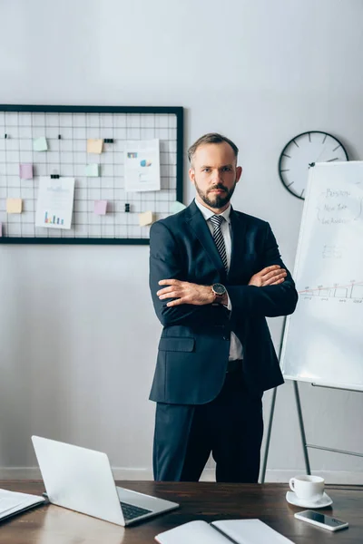 Serious businessman looking at camera with crossed arms near devices and notebook on blurred foreground — Stock Photo