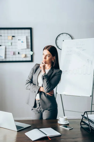 Donna d'affari pensierosa in piedi vicino a dispositivi e notebook in primo piano sfocato in ufficio — Foto stock