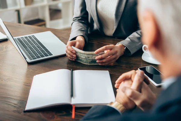 Cropped view of businesswoman holding money near laptop, notebook and businessman in blurred foreground — Stock Photo