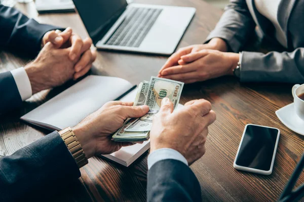 Vista recortada del hombre de negocios sosteniendo dinero cerca de teléfono inteligente, café y colegas en un fondo borroso - foto de stock