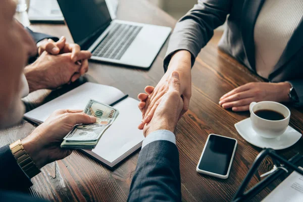 Businessman holding money and shaking hands with colleague near smartphone and coffee on table — Stock Photo