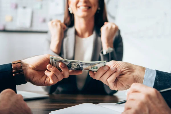 Vista recortada de los hombres de negocios sosteniendo el dinero cerca de la mujer de negocios mostrando sí gesto sobre fondo borroso - foto de stock