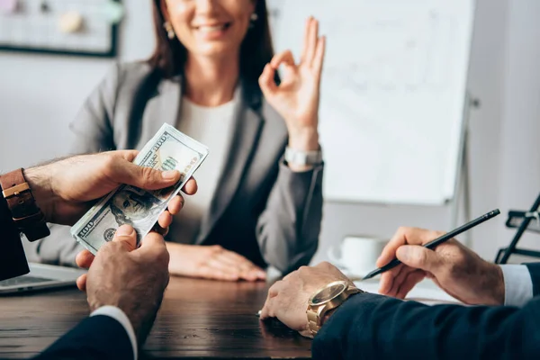 Vista recortada de hombre de negocios sosteniendo dinero en efectivo cerca de colega con pluma y mujer de negocios mostrando bien en fondo borroso - foto de stock