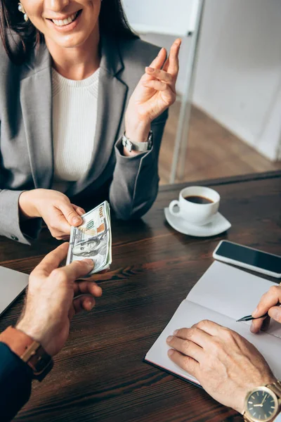 Cropped view of businessman giving money to smiling businesswoman near colleague with notebook — Stock Photo