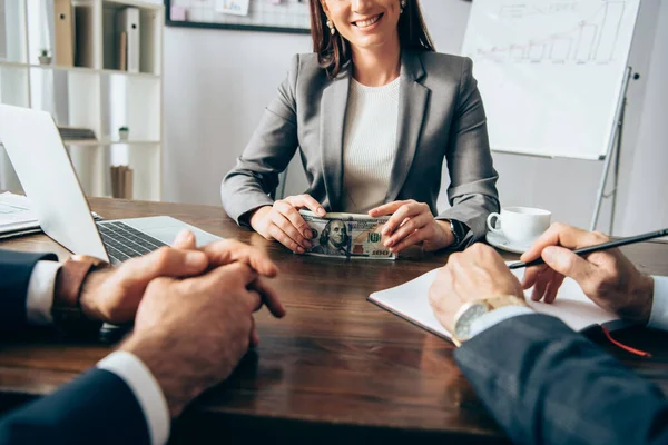 Vue recadrée d'une femme d'affaires souriante tenant des dollars près d'un ordinateur portable et de collègues au premier plan flou au bureau — Photo de stock