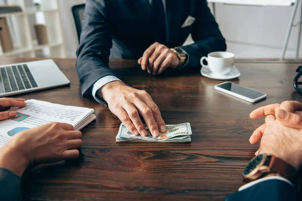 Cropped view of businessman holding dollars near devices and colleagues with documents on blurred foreground — Stock Photo