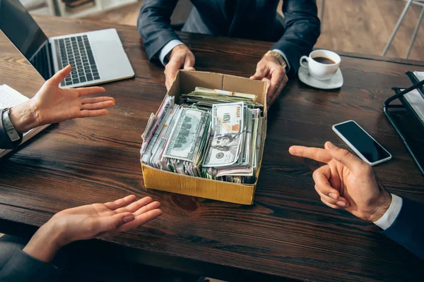 Cropped view of businesspeople pointing at box with money near colleague and devices on blurred foreground — Stock Photo