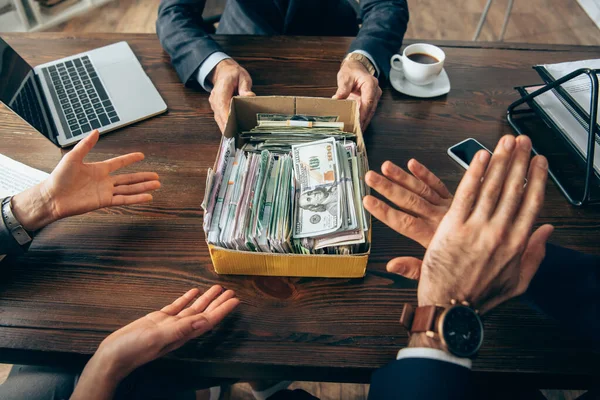 Cropped view of businessman holding box with cash near colleagues showing stop gesture on blurred foreground — Stock Photo