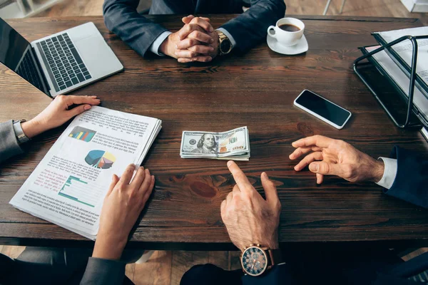 Cropped view of businessman pointing at money near colleague with papers and devices on table — Stock Photo