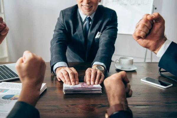 Vista recortada del inversor sonriente sosteniendo efectivo cerca de colegas mostrando sí gesto en primer plano borroso en la oficina - foto de stock