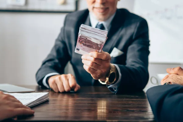Cropped view of investor holding money near businesspeople with documents on blurred foreground — Stock Photo
