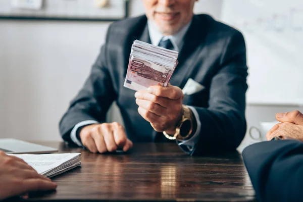 Vista recortada del inversor sosteniendo dinero cerca de los empresarios con documentos en primer plano borroso - foto de stock