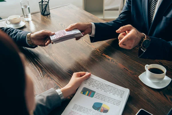 Inversor dando dinero a hombre de negocios y apuntando a mujer de negocios con papeles en primer plano borrosa - foto de stock