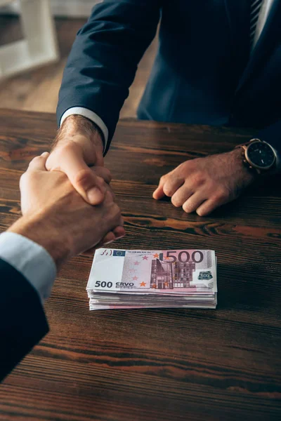 Vista recortada de homens de negócios a apertar as mãos perto das notas de euro na mesa — Fotografia de Stock