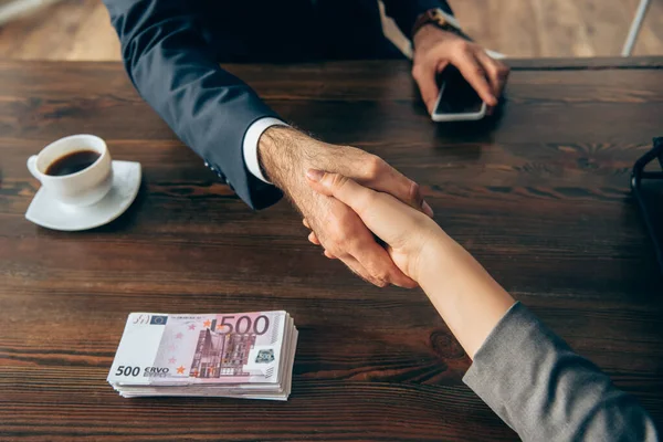Vista ritagliata dei partner commerciali che stringono la mano vicino al caffè e alle banconote in euro sul tavolo — Foto stock