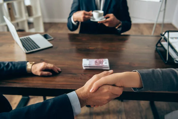 Vista recortada de los socios de negocios estrechando las manos cerca del dinero y colega con taza sobre fondo borroso — Stock Photo