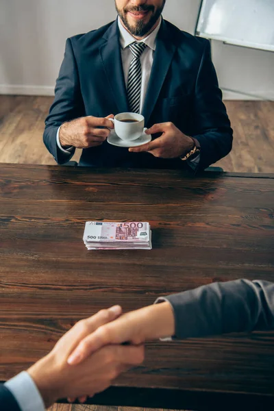 Cropped view of businessman holding cup of coffee near money and colleagues handshaking on blurred foreground — Stock Photo