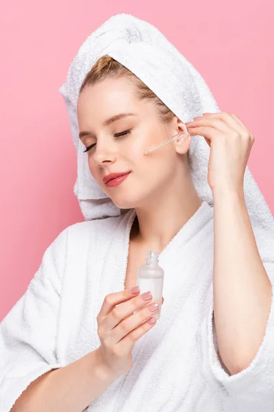 Mujer joven en albornoz con toalla en la cabeza aplicando suero en la cara aislado en rosa — Stock Photo